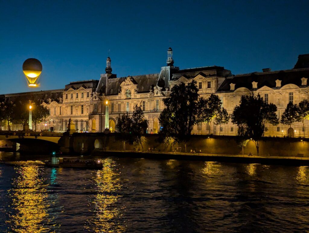 Photograph of the Seine at night with the Olympic balloon rising over the buildings in the top left of the shot. 